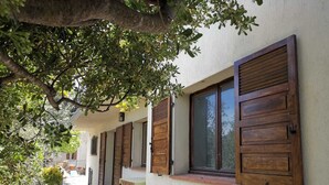 A row of rooms with wooden shutters, a stone-paved walkway, and a tree providing shade. - Bas de Villa au Calme (Villeneuve-Loubet)