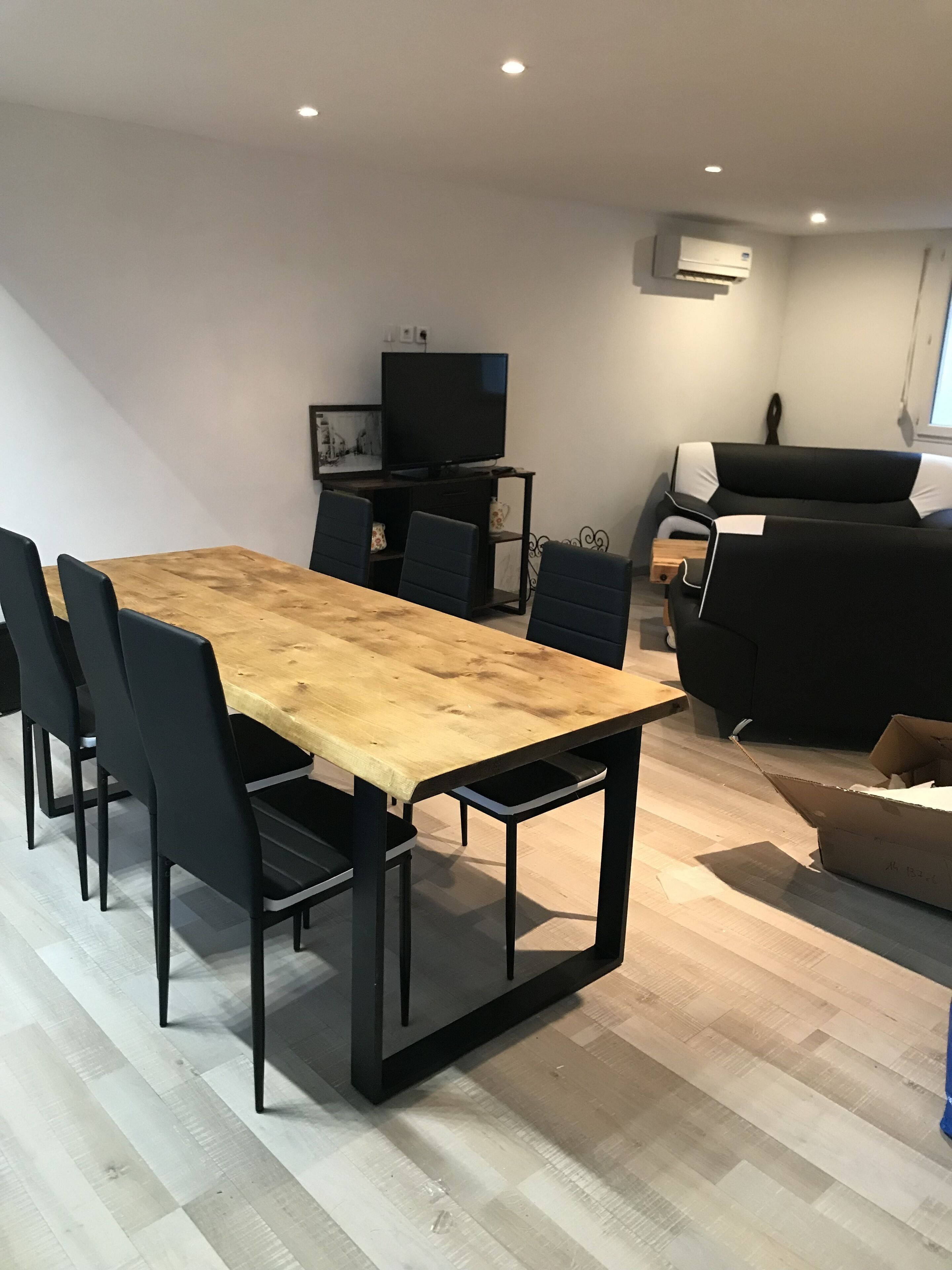 Una sala de estar moderna con una mesa de comedor de madera, sillas negras, un sofá negro, un televisor sobre un mueble bajo y una unidad de aire acondicionado en la pared.