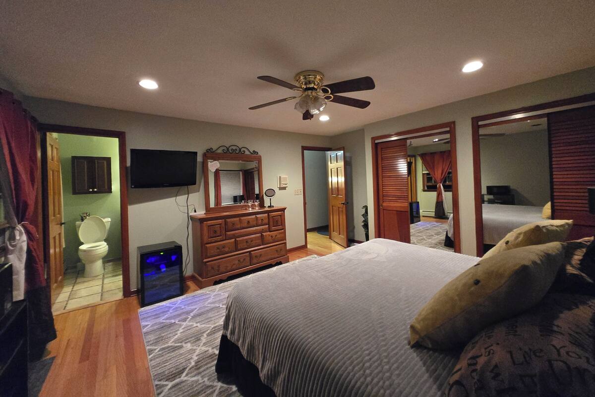 A bedroom with a bed, dresser, ceiling fan, and a television on the wall.