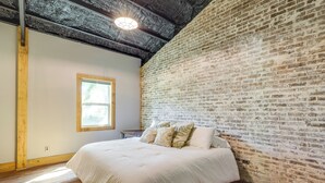 A bedroom with a brick wall, a wooden floor, a large bed with white bedding, and a window with a view. - Fish & Relax! Magnolia River Cabin Hideaway (Magnolia Springs)