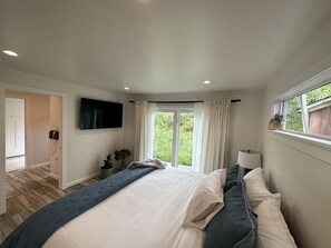 Une chambre à coucher avec un lit, un téléviseur fixé au mur, une fenêtre avec des rideaux et une vue sur la verdure à l’extérieur.