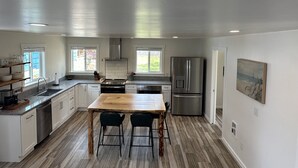 Une cuisine moderne avec une table à manger en bois, deux tabourets de bar, des appareils électroménagers en acier inoxydable et une vue sur le jardin à travers de grandes fenêtres.