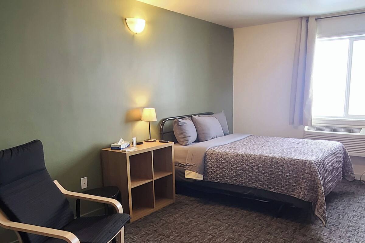 A hotel room with a bed, a chair, a bedside table with a lamp, and a window with curtains.