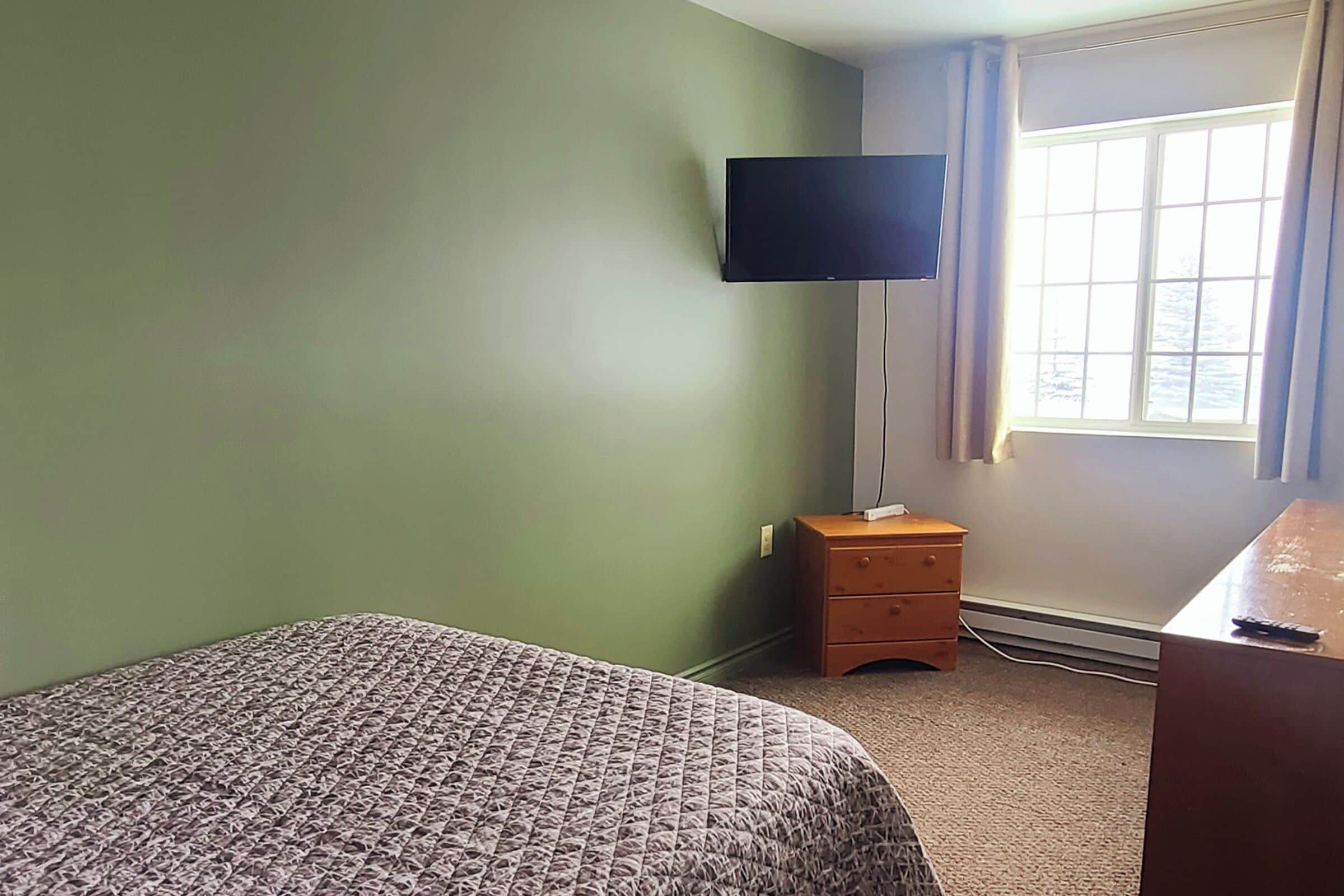 A room with a bed, a television mounted on the wall, a small wooden nightstand, and a window with curtains.