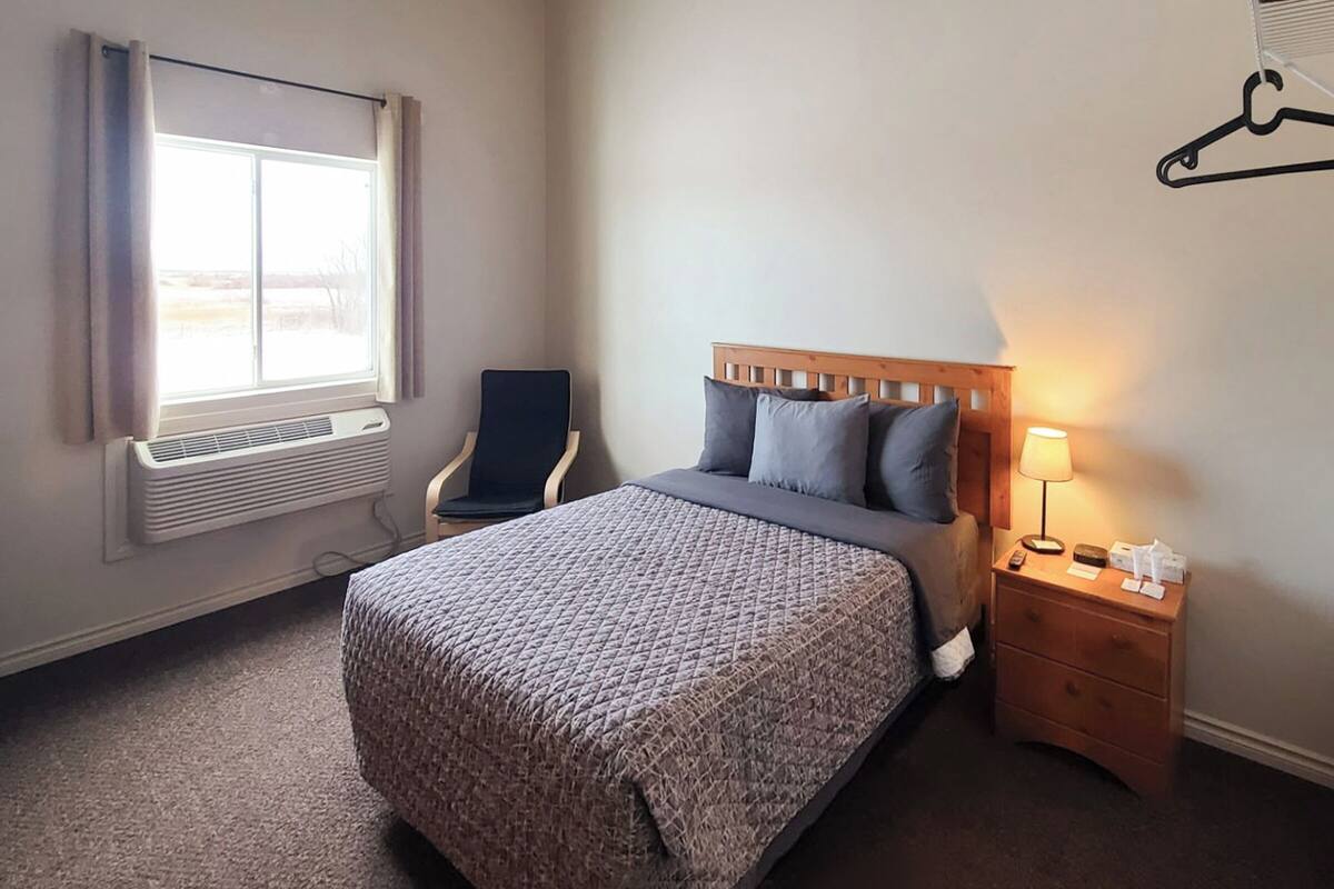 A hotel room with a bed, a chair, a bedside table with a lamp, and a window with curtains.