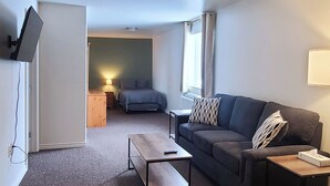 Een knus woongedeelte met een bank, een salontafel, een aan de muur bevestigde televisie en een raam met gordijnen.