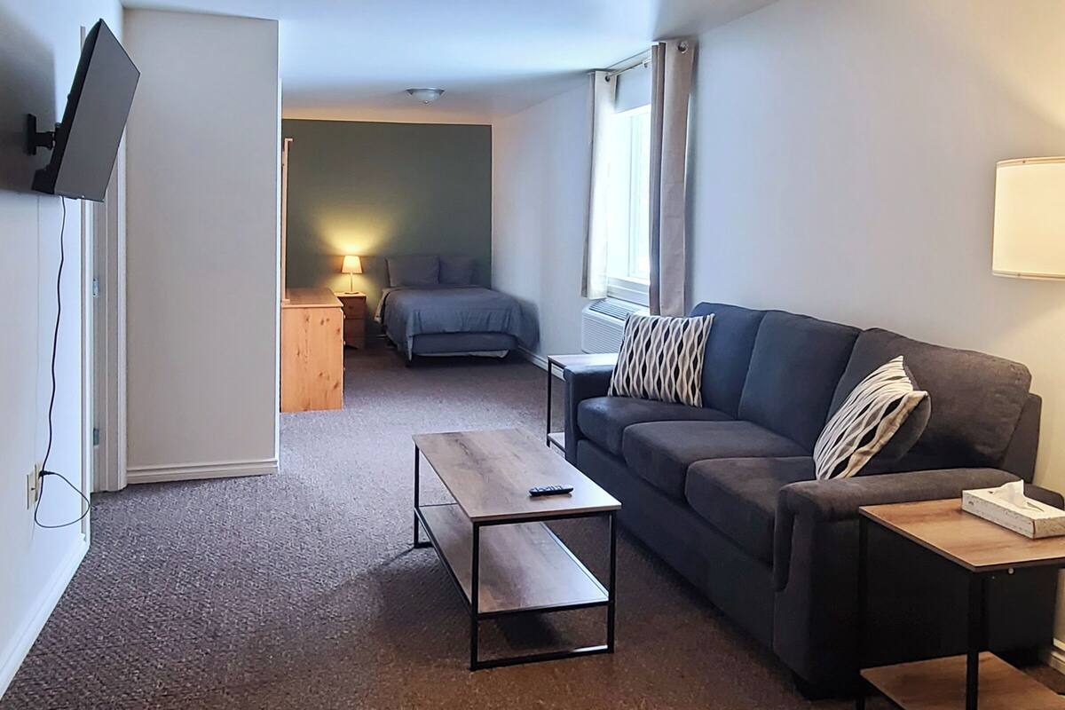 A compact living space with a sofa, a coffee table, a TV mounted on the wall, and a window with curtains.
