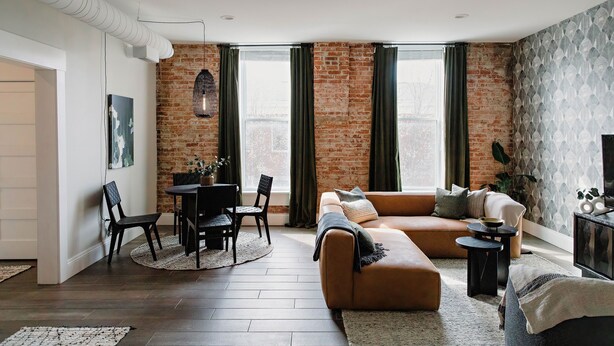 Una sala moderna con pared de ladrillo, un sofá de cuero marrón, un comedor con mesa y sillas, y un televisor sobre un mueble bajo.