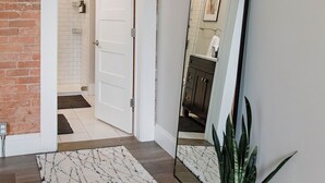Un pasillo con pared de ladrillo, una puerta blanca, un espejo, un tapete estampado y una planta en maceta.