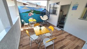 Un balcón con piso de madera, sillas amarillas y blancas, una mecedora azul y una mesa con un florero.