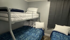Ein kleines Hotelzimmer mit einem Einzelbett und zwei Etagenbetten, einem Fenster mit Vorhang und einer Deckenleuchte.