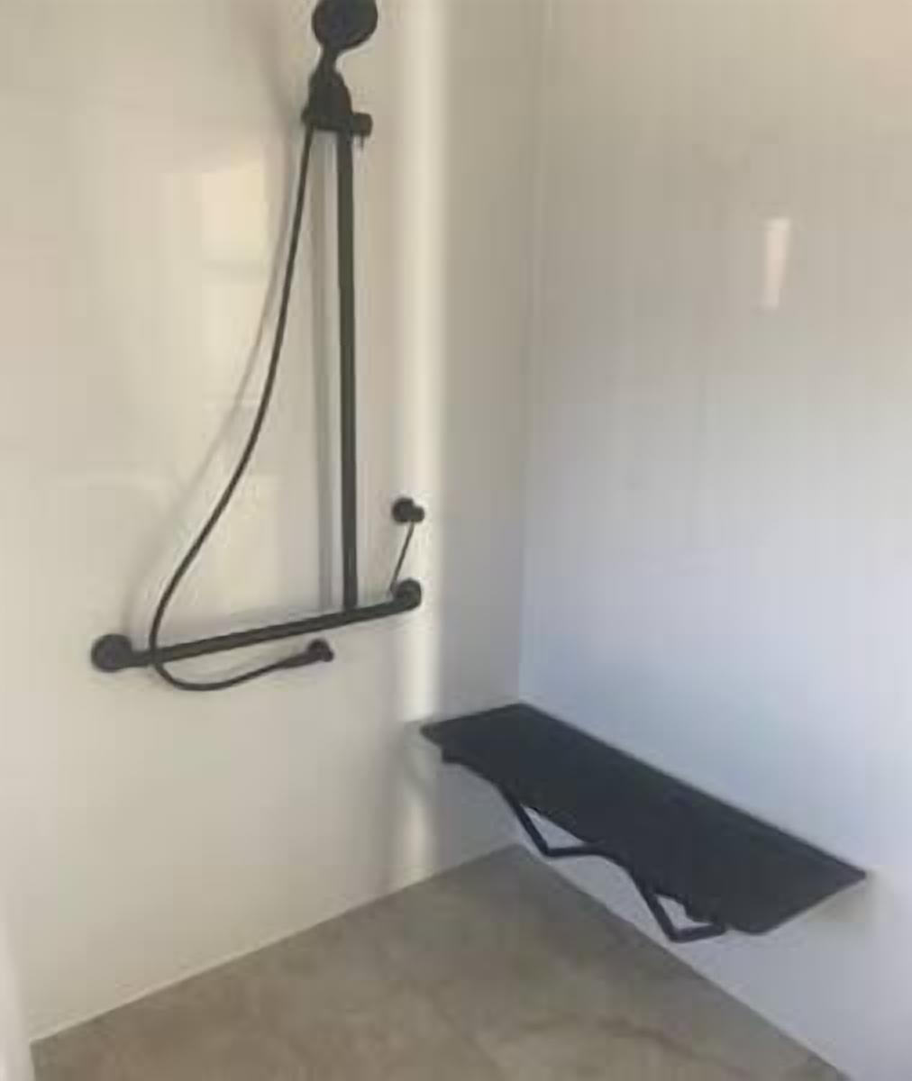 Un coin d’une pièce comprenant un espace douche, avec un pommeau de douche, une étagère fixée au mur et un sol carrelé.