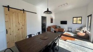 A spacious living area with a wooden dining table, black chairs, a sofa, and a TV mounted on the wall.