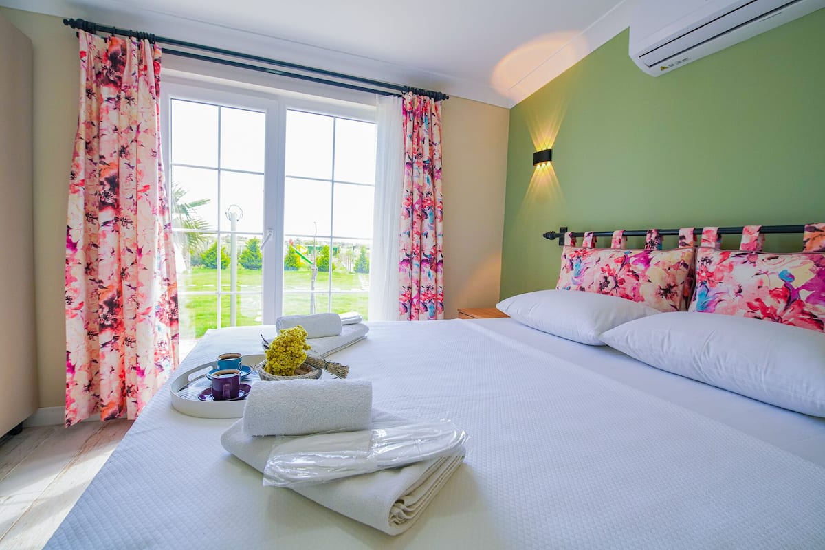 A hotel room with a bed, a tray with towels and a cup, and a view of a green outdoor area through a window.