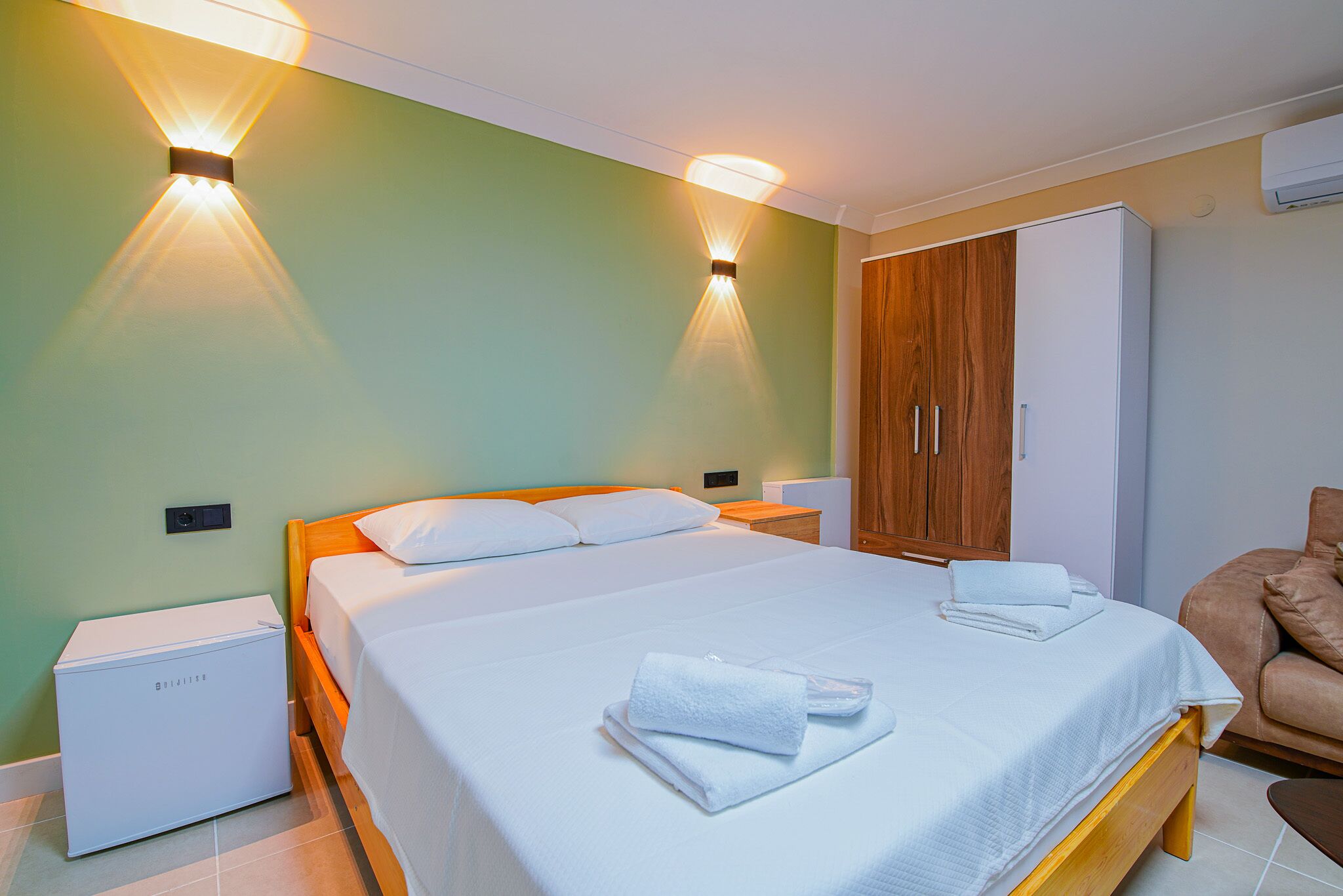Een keurig opgemaakt bed met witte lakens en handdoeken, een houten nachtkastje met een wit apparaat, een houten garderobe en een leren fauteuil.