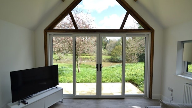 Una habitación con un gran ventanal corredero que da a un jardín, un televisor de pantalla plana sobre un aparador blanco y una ventana con vistas a árboles.
