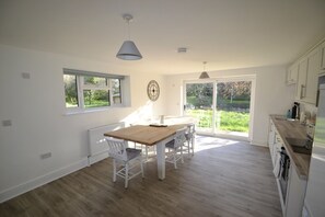 Una cocina luminosa con una mesa y sillas de madera, armarios blancos y vistas a la vegetación a través de las ventanas.
