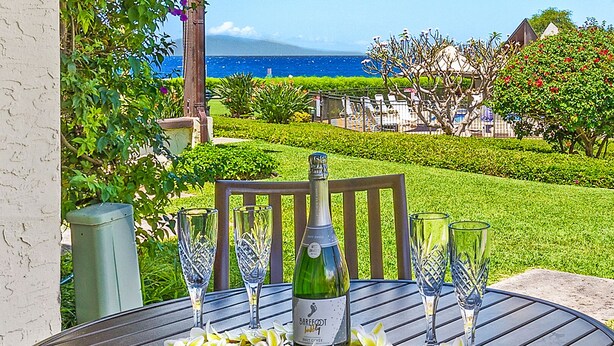 A table set outdoors with a bottle and glasses, surrounded by greenery and a view of the ocean. - Hale Kamaole #166, VIEW, No Steps to Entry, Pool, A+ Wi-Fi, King Bed, EZ Parking (Kihei)