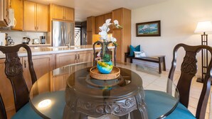 A dining area with a glass table, blue cushioned chairs, a vase with flowers, and wooden cabinets. - Hale Kamaole #166, VIEW, No Steps to Entry, Pool, A+ Wi-Fi, King Bed, EZ Parking (Kihei)