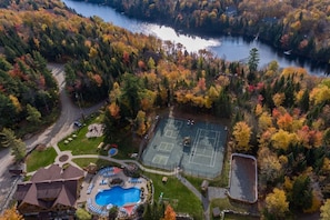 An aerial view of a property with a tennis court, swimming pool, and a lake surrounded by autumn-colored trees.