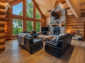 A log cabin interior with a stone fireplace, leather sofas, and a large window overlooking greenery.