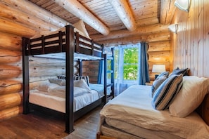 A log cabin room with a bunk bed and a single bed, wooden walls, and a window with a view of greenery.