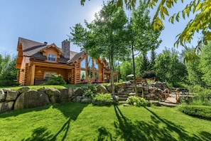 A log cabin with a stone chimney, a stone retaining wall, and a well-maintained lawn.