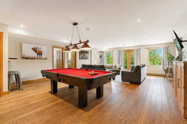 A pool table in a room with a sofa, a television, and large windows.