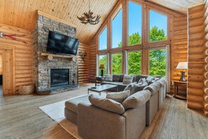 A log cabin interior with a stone fireplace, a large TV, a sofa, and large windows.