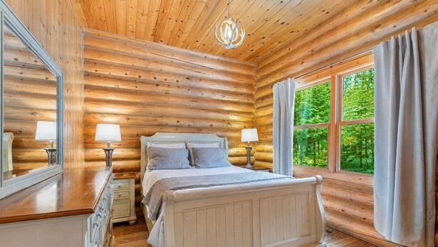 A log cabin bedroom with a bed, bedside tables, and a large window with curtains. - Fiddler Lake Resort - Elk 96 (Mille-Isles)
