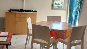 A room with a dining table, chairs, a TV mounted on the wall, a window with curtains, and a wooden cabinet. - Meublé de Tourisme 3 Étoiles en Résidence Sécurisé (Cap d'Agde)