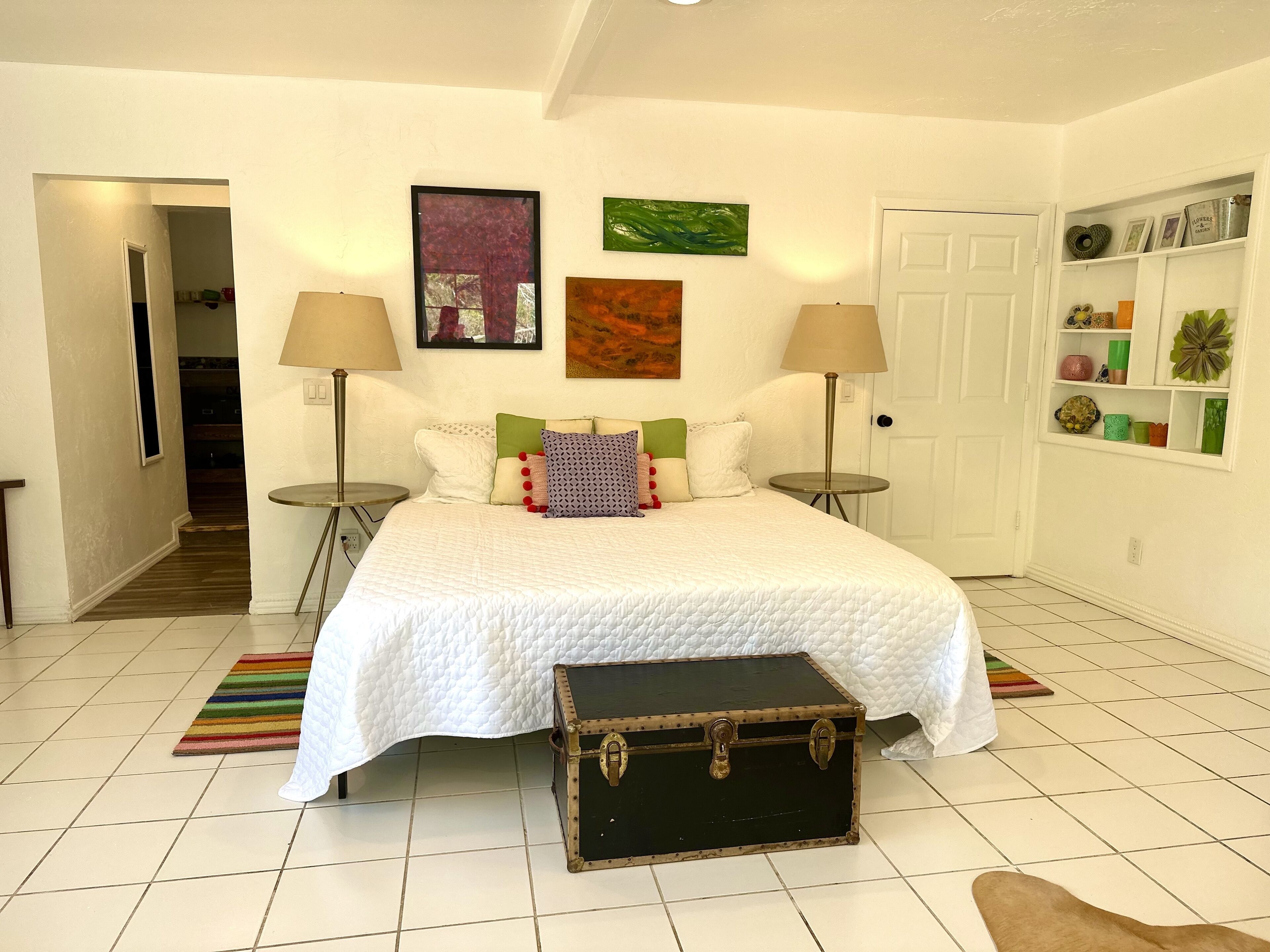 Een slaapkamer met een bed, twee bedlampen, een koffer, een kleurrijk vloerkleed en een deur naar een hal.