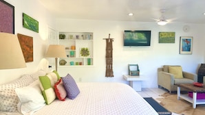 Une pièce bien éclairée, avec un lit, un téléviseur fixé au mur, un ventilateur de plafond et une petite table avec une chaise.