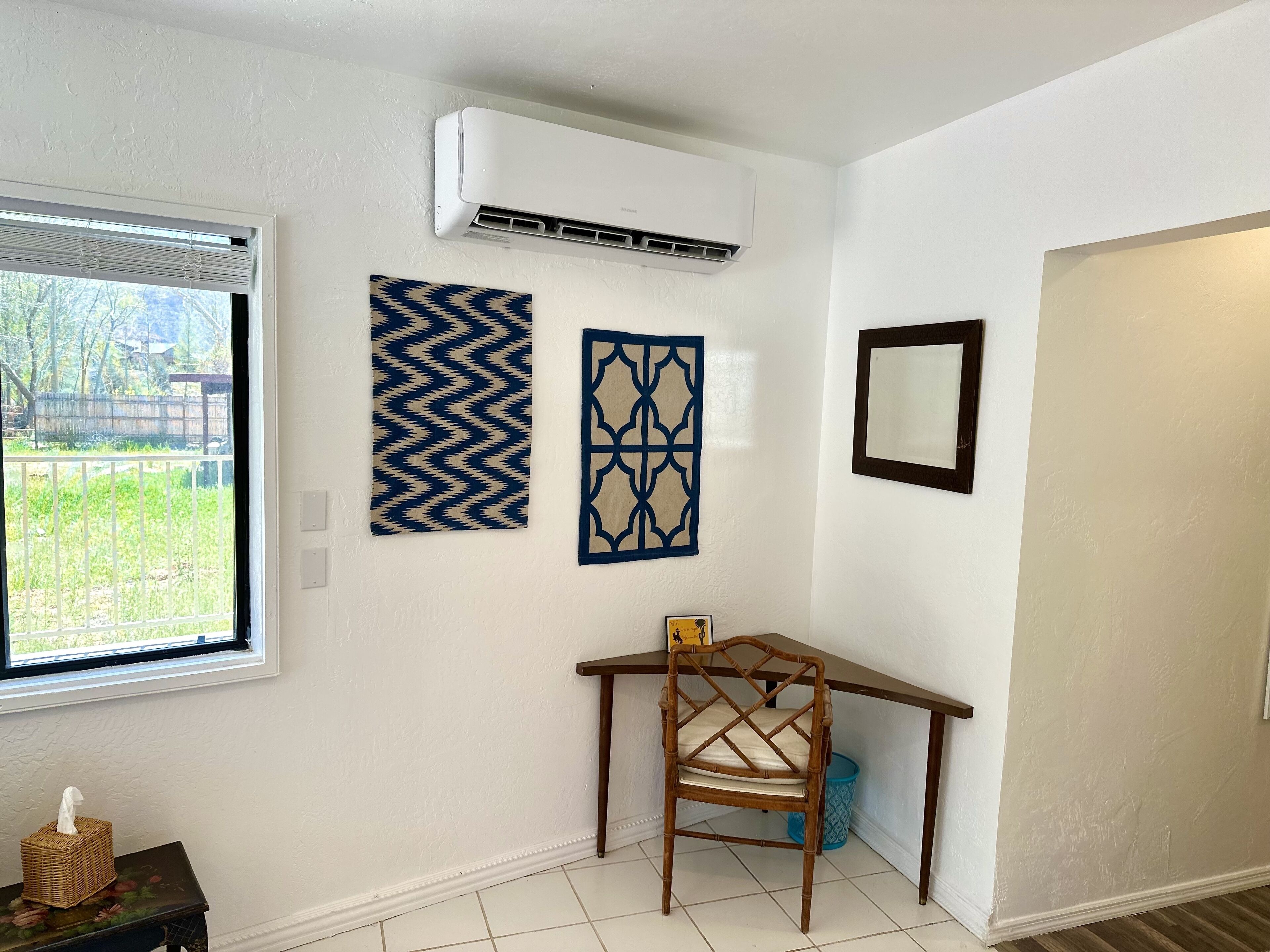 A room with a window, an air conditioner, a chair, a small table, and a wall with two decorative panels.