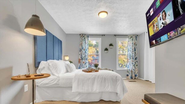 Une chambre à coucher avec un lit, une table de chevet, une lampe, une télévision et des fenêtres avec des rideaux.
