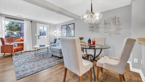 Un salon moderne avec un coin repas, comprenant une table en verre, des chaises blanches et un tapis à motifs.