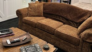 A cozy living room with a patterned sofa, a wooden coffee table with books and a remote control, and a kitchen area in the background. - Your Very Own Sports Bar! (Post Falls)