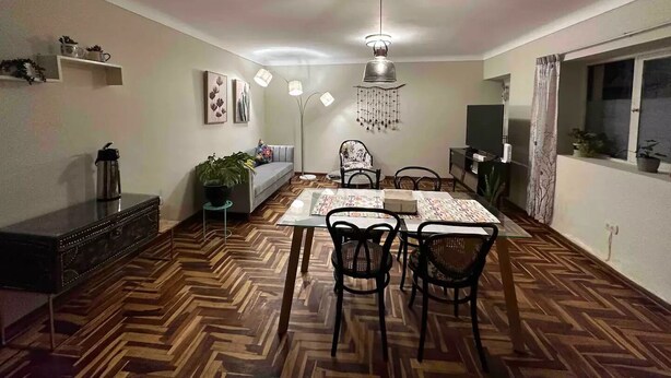 Un comedor con piso de madera en espiga, una mesa central con sillas, una lámpara colgante y un televisor a la derecha.
