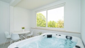 Un baño moderno con hidromasaje, paredes blancas, un ventanal con vistas a árboles y una pequeña mesa con sillas.