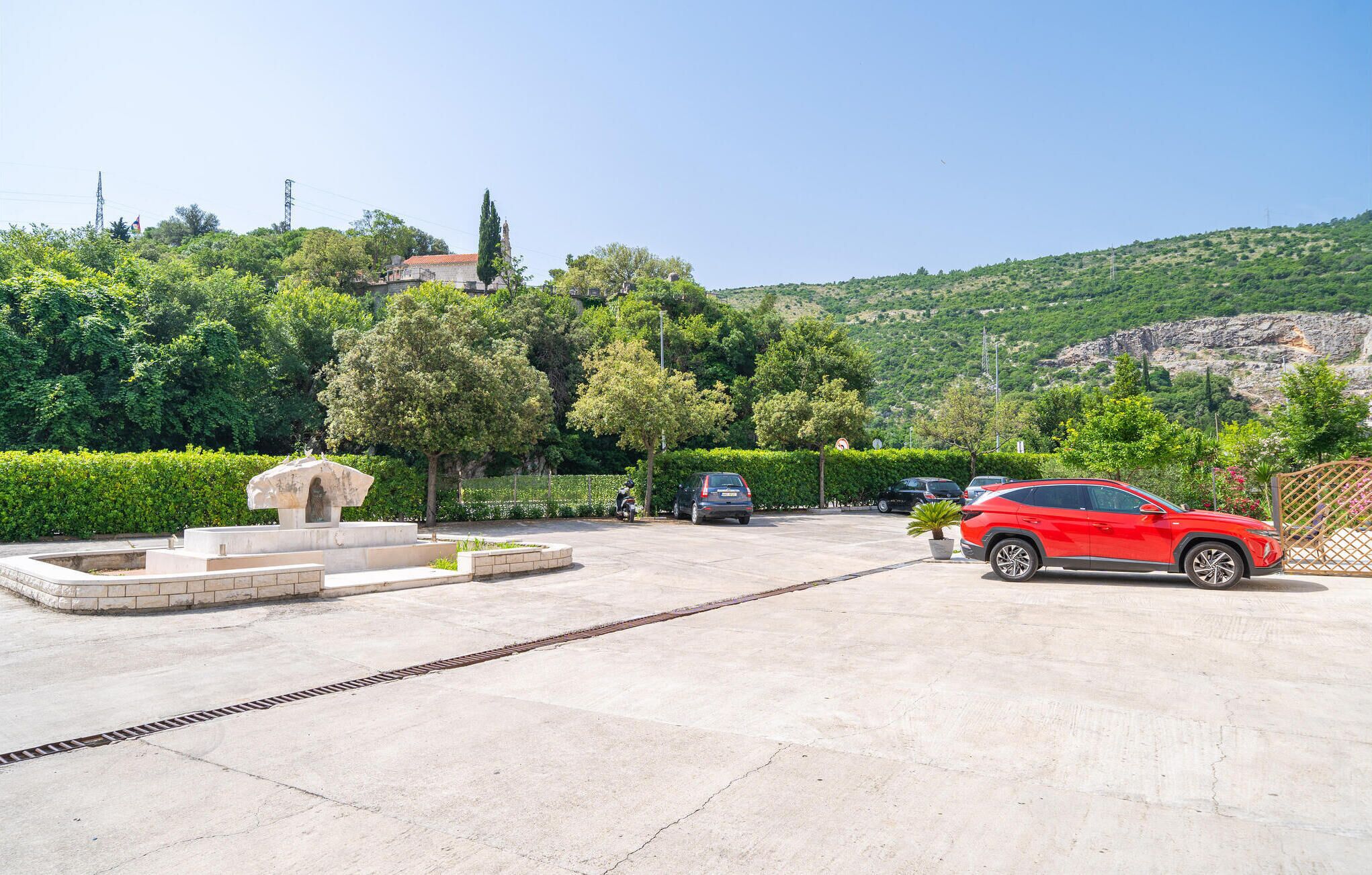 Un espace de stationnement avec une voiture rouge, un monument en pierre et une vue sur une colline parsemée d’arbres, avec un bâtiment visible au loin.