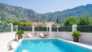 Una piscina con vistas a montañas, un patio con sillones de descanso y una casa al fondo.