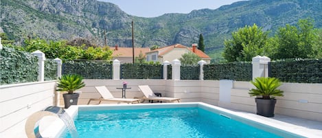 Ein Schwimmbad mit Bergblick, eine Terrasse mit Liegestühlen und ein Haus im Hintergrund.