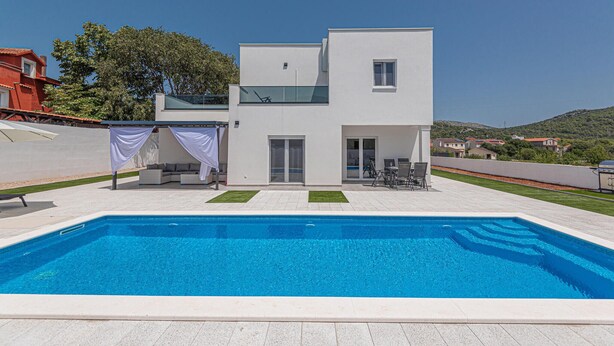 Ett modernt hus i två våningar med en swimmingpool, en täckt uteplats och utsikt över det omgivande landskapet.