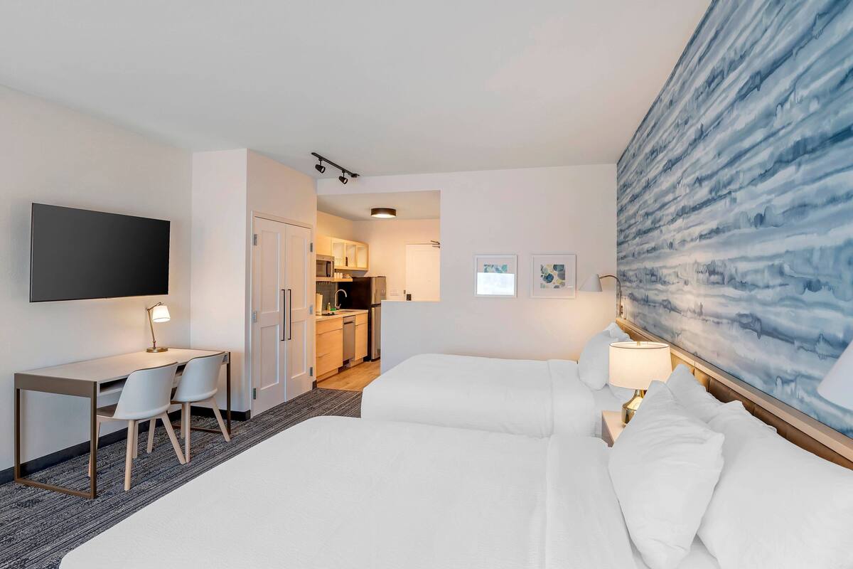 Une chambre d’hôtel avec un lit, un bureau, des chaises, une télévision, un tableau et une vue sur la cuisine.