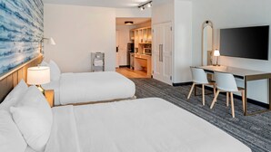 Ein Hotelzimmer mit zwei Betten, einem Schreibtisch mit Stühlen, einem Fernseher und einer Küche im Hintergrund.
