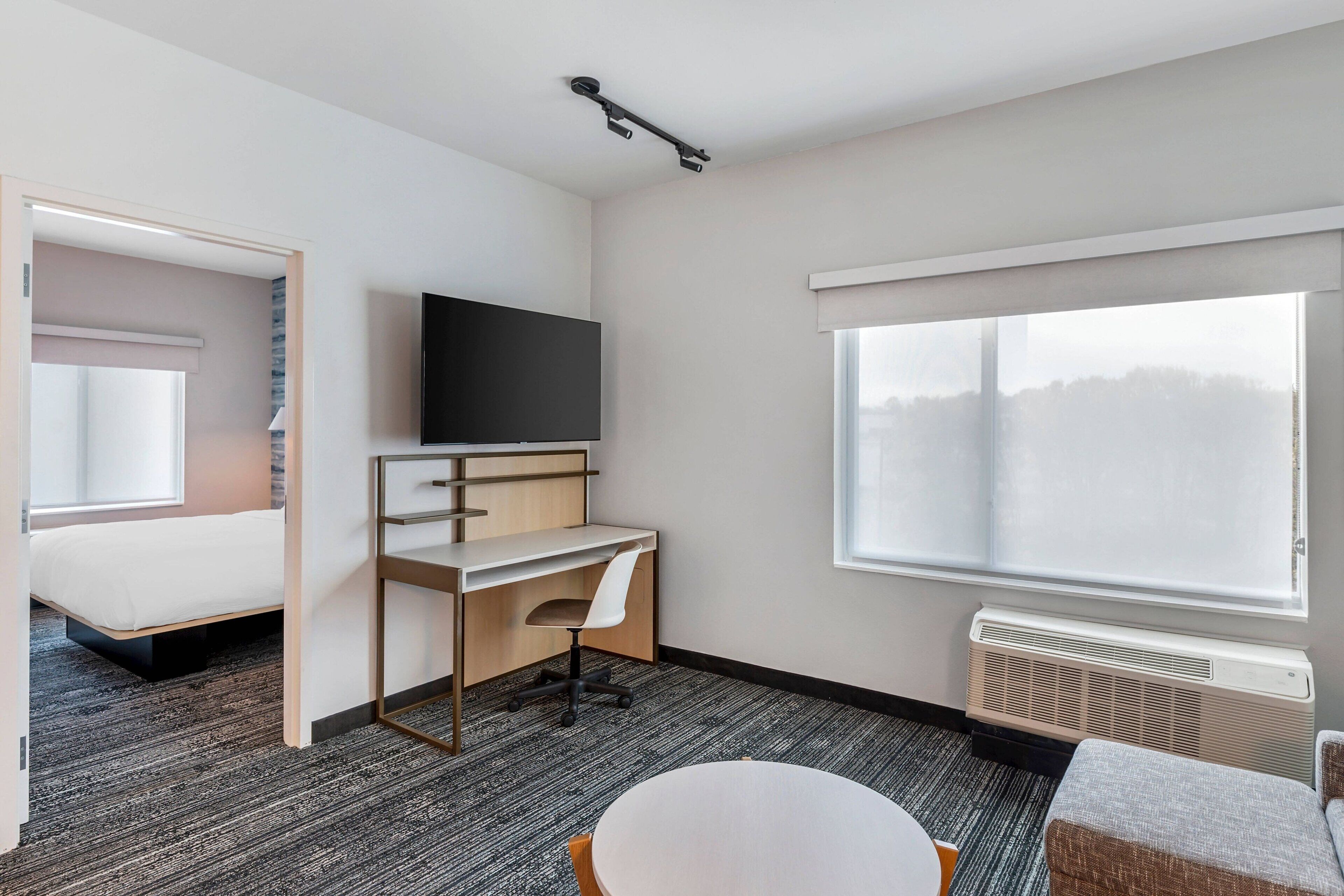 A modern hotel room with a bed, a desk with a chair, a television, a round table, and a large window.