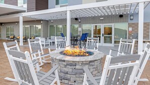 Eine Terrasse mit einem Steinkamin, umgeben von weißen Holzstühlen, einem überdachten Sitzbereich und einem Gebäude mit großen Fenstern und Außenplätzen.