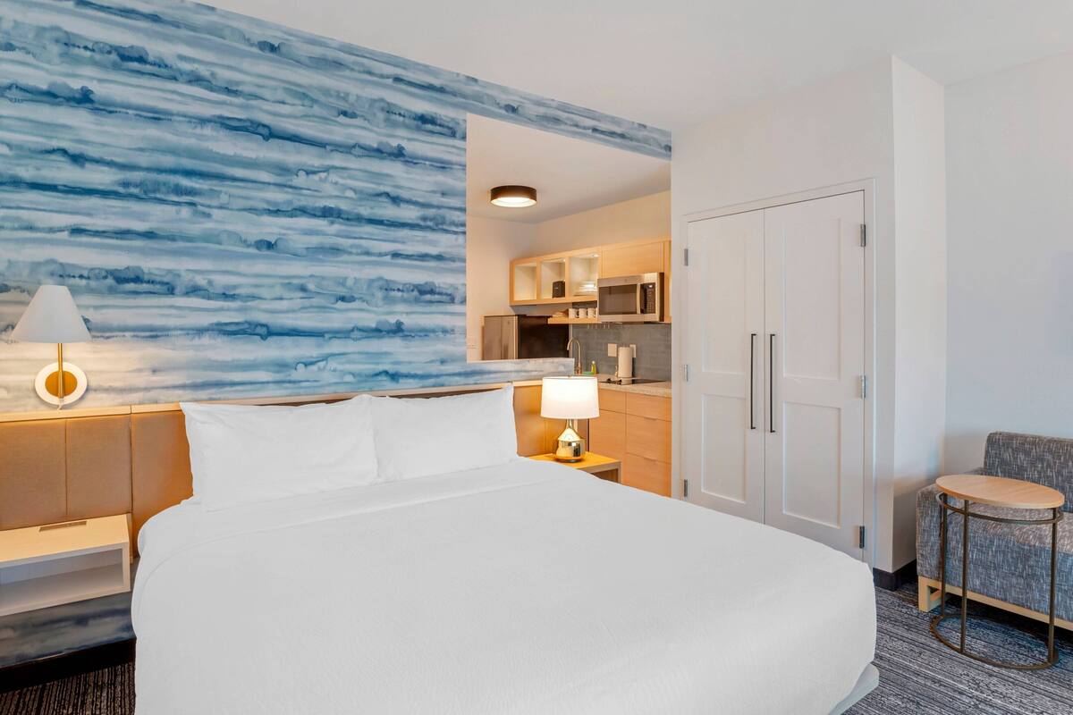 Une chambre d’hôtel avec un grand lit, une table de chevet avec une lampe, un coin salon avec une petite table et, au fond, une kitchenette.