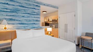 A hotel room with a large bed, a nightstand with a lamp, a seating area with a small table, and a kitchenette in the background.