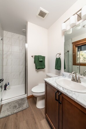 A modern bathroom with a glass shower enclosure, a toilet, a sink with a marble countertop, and wooden cabinets. - Evergreen Cabin Rental (Valemount)
